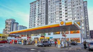 A Shell gas station with multiple fuel pumps, cars refueling, and a Shell Select store, set against tall modern apartments