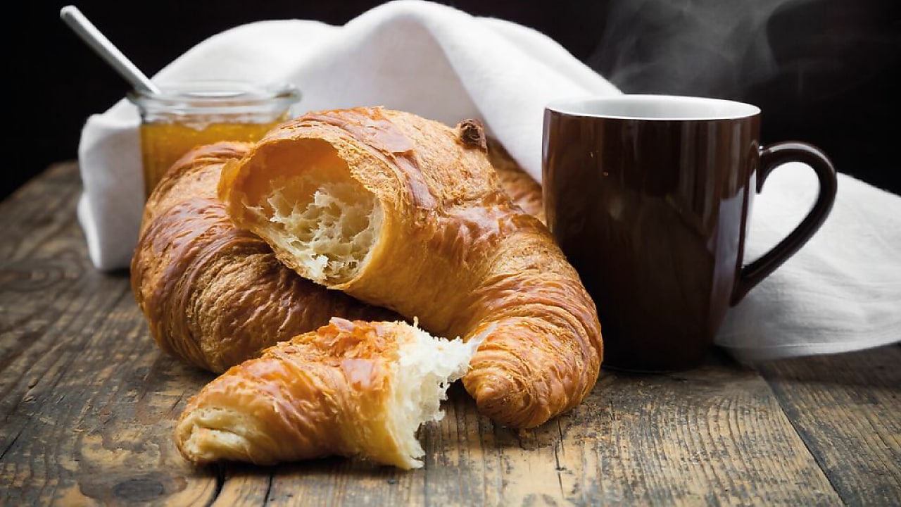 A steaming cup of coffee with golden, flaky croissants on a rustic wooden table at Artcaffé. A jar of jam sits in the background.