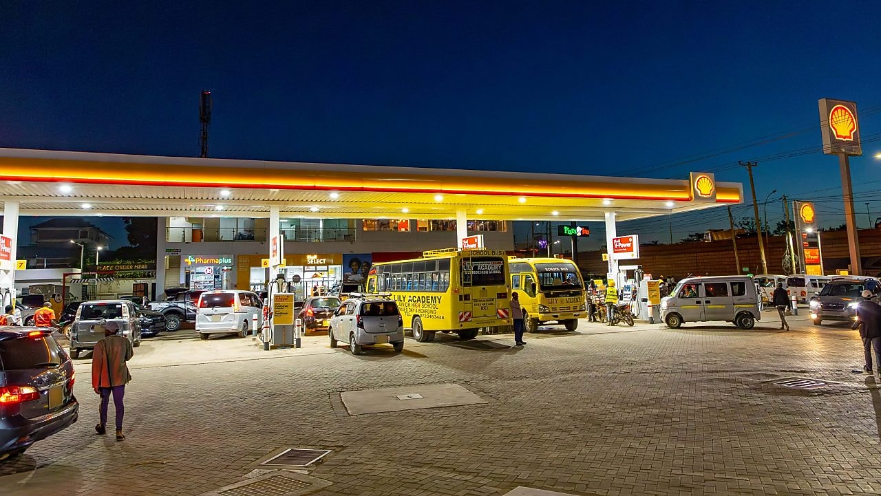 A busy Shell gas station at night with school buses, cars, and motorcycles refueling. A convenience store and pharmacy are in the background, with people walking and waiting