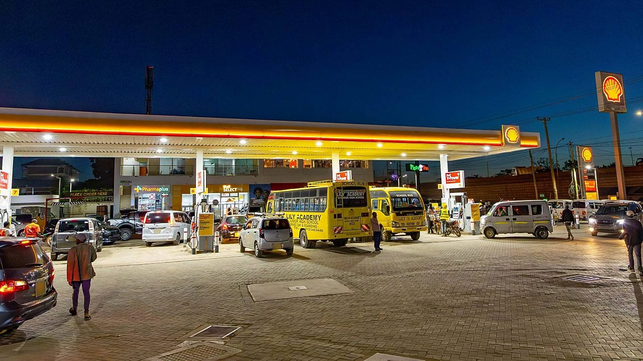 A busy Shell gas station at night with school buses, cars, and motorcycles refueling. A convenience store and pharmacy are in the background, with people walking and waiting