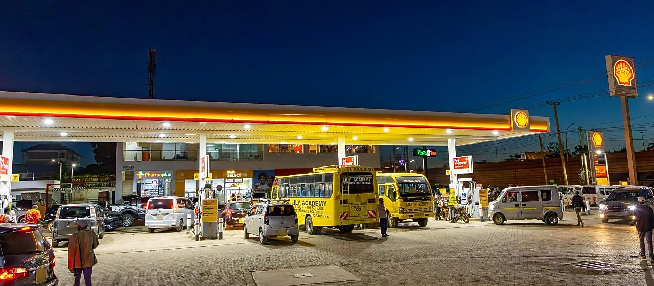 A busy Shell gas station at night with school buses, cars, and motorcycles refueling. A convenience store and pharmacy are in the background, with people walking and waiting