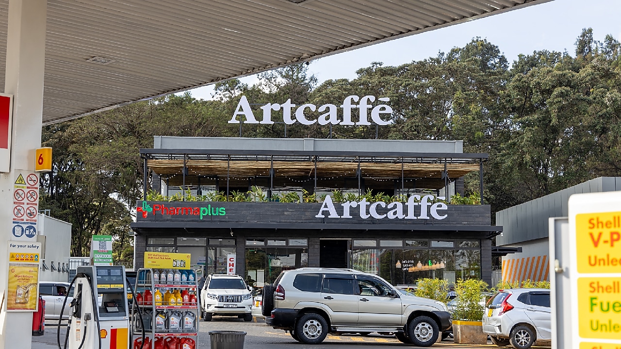 A Shell fuel station with fuel pumps in the foreground and an Artcaffé café above a PharmaPlus pharmacy in the background. Cars are parked and moving around the forecourt.