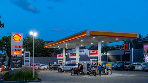 A busy Shell fuel station in the evening with customers on the forecourt, cars and motorcycles refueling, and a convenience store