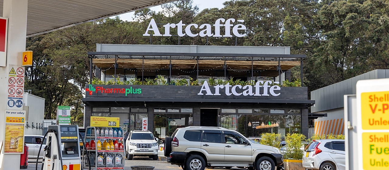 A Shell fuel station with fuel pumps in the foreground and an Artcaffé café above a PharmaPlus pharmacy in the background. Cars are parked and moving around the forecourt.