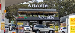 A Shell fuel station with fuel pumps in the foreground and an Artcaffé café above a PharmaPlus pharmacy in the background. Cars are parked and moving around the forecourt.