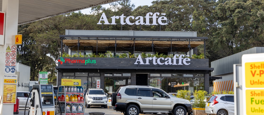 A Shell fuel station with fuel pumps in the foreground and an Artcaffé café above a PharmaPlus pharmacy in the background. Cars are parked and moving around the forecourt.