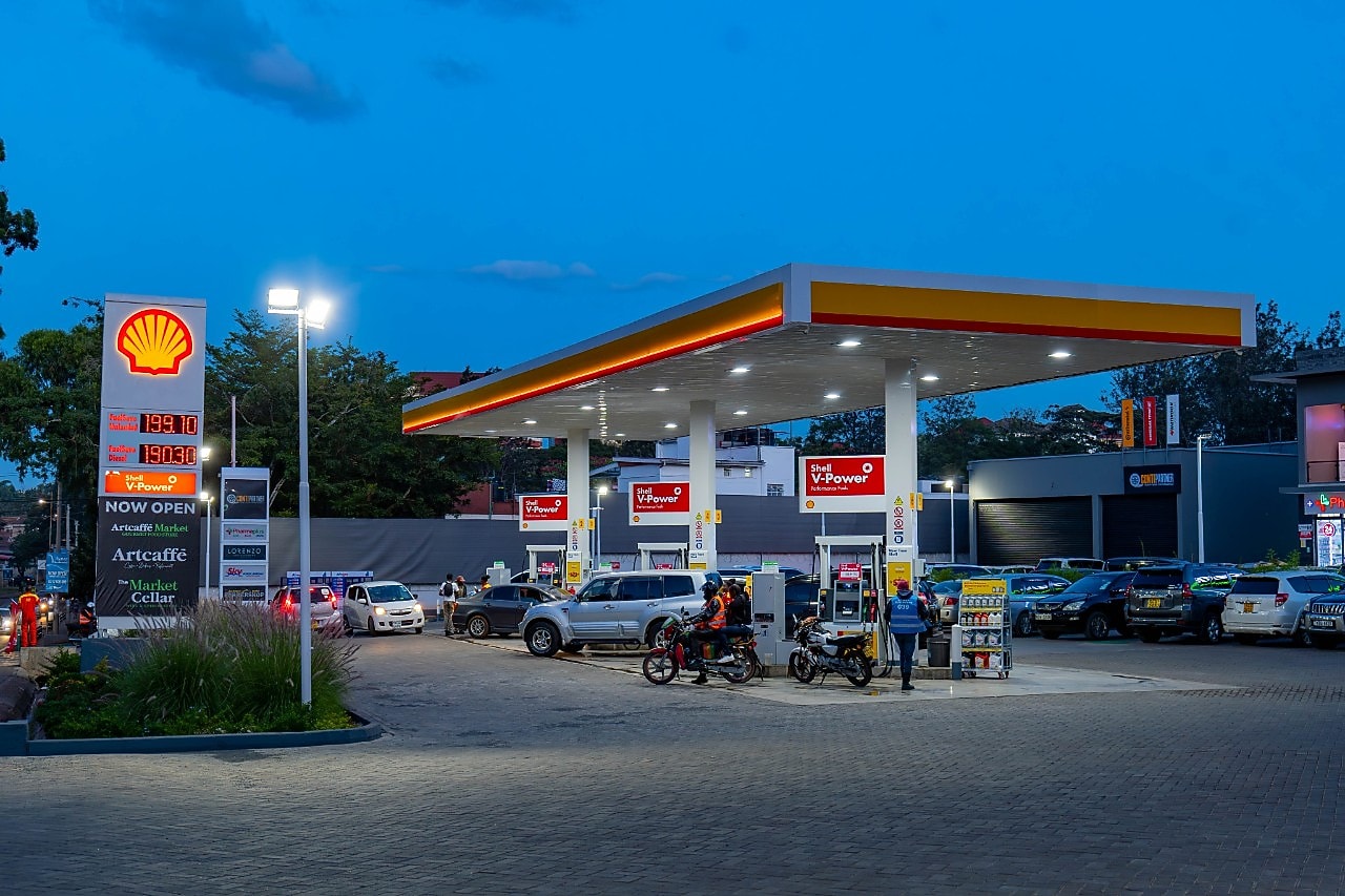 A busy Shell gas station at night with school buses, cars, and motorcycles refueling. A convenience store and pharmacy are in the background, with people walking and waiting
