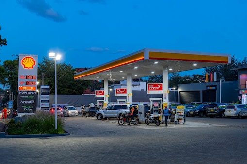 A busy Shell gas station at night with school buses, cars, and motorcycles refueling. A convenience store and pharmacy are in the background, with people walking and waiting